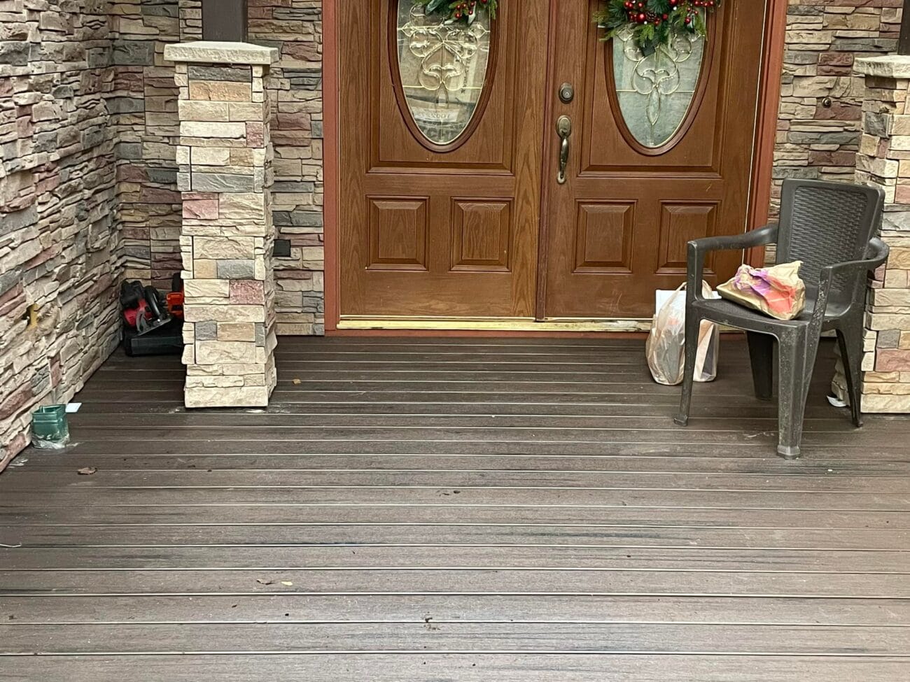 Front porch with dark wooden flooring, two double wooden doors, a plastic chair, a plastic bag, a drink cup, and a black tray near a stone column.