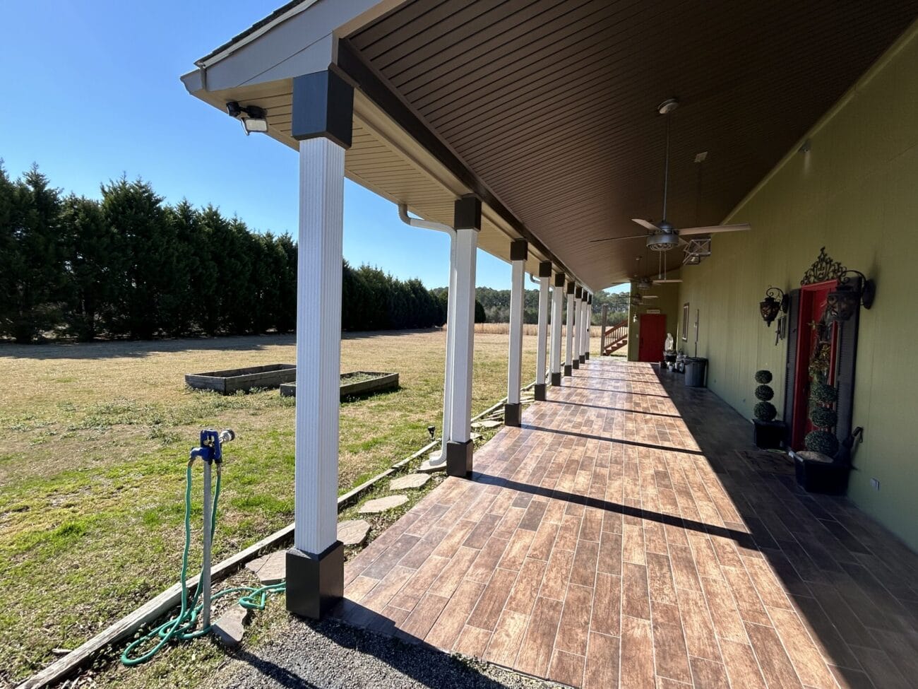 A covered patio with tiled flooring, white columns, ceiling fans, and a red door overlooks a grassy yard with a garden bed and hose in the foreground.