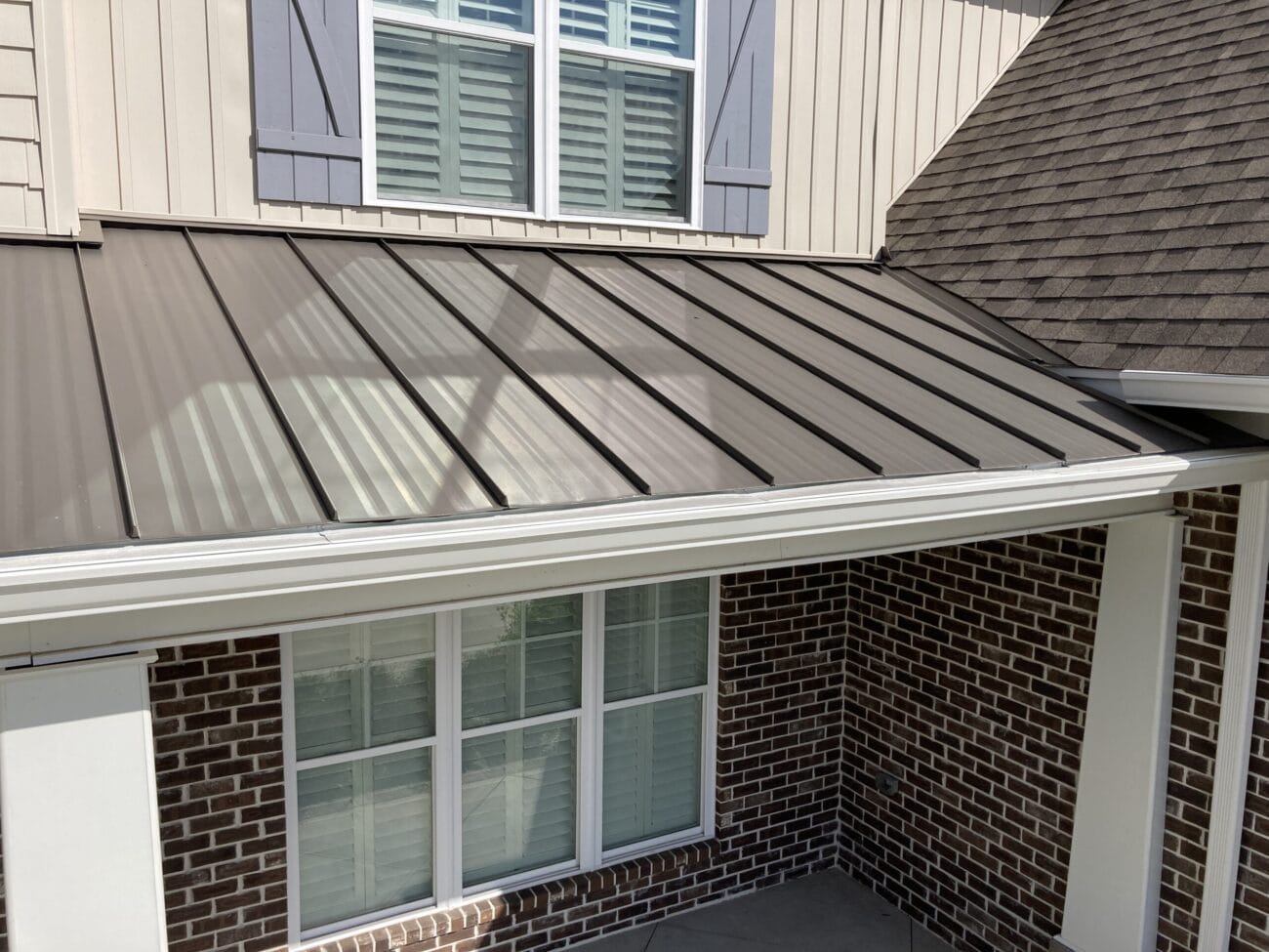 A close-up view of a house exterior showing a standing seam metal roof above brick walls and windows with white shutters.