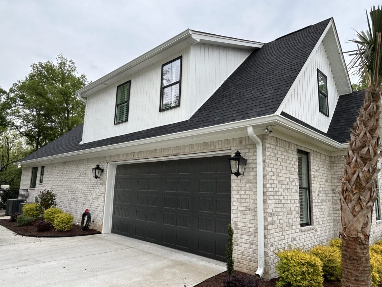 A two-story house with white brick exterior, black garage door, black roof, and landscaped yard with shrubs and a palm tree.