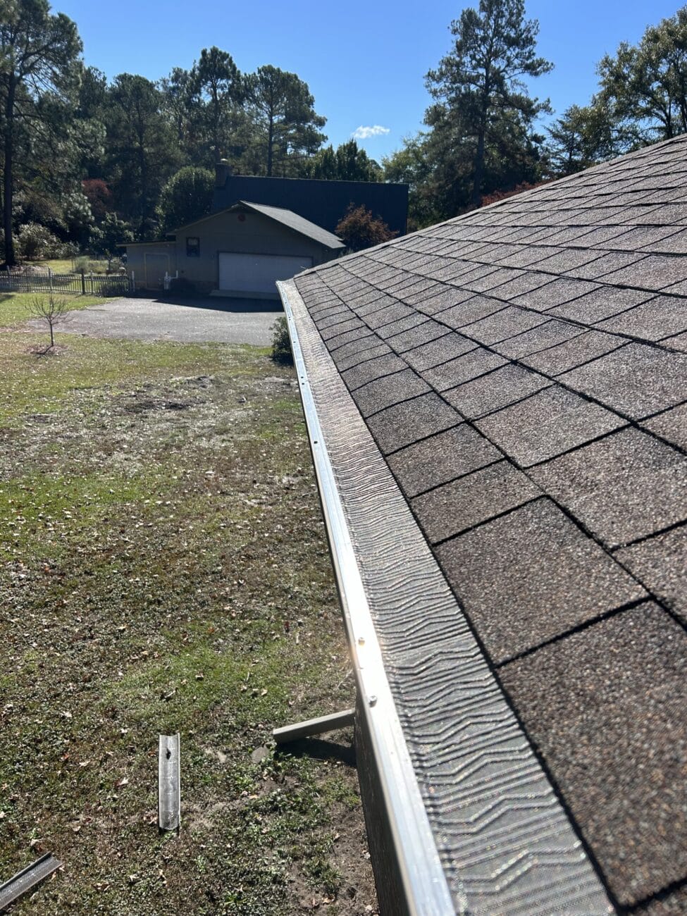 A view from a rooftop showing an asphalt shingle roof and a metal gutter guard, with a grassy yard, trees, and a detached garage in the background.