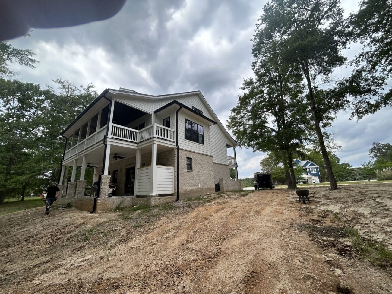 Two-story white house with wraparound porch sits on a sloped, partially cleared lot under a cloudy sky; a person stands to the left and a blue house is visible in the background.