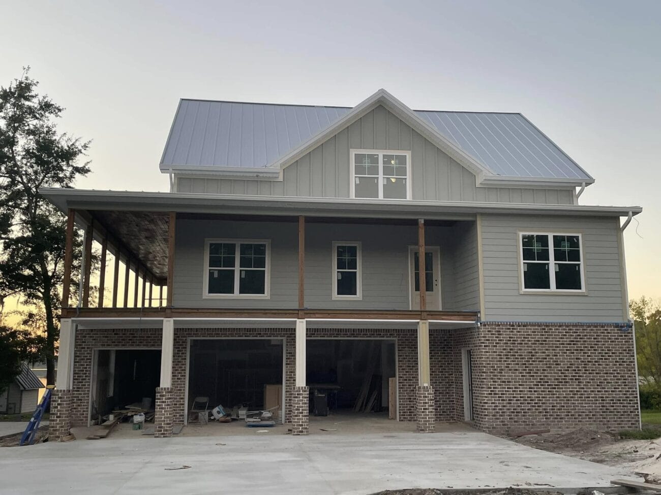 A two-story house under construction with gray siding, brick foundation, large garage doors, and an unfinished porch area.