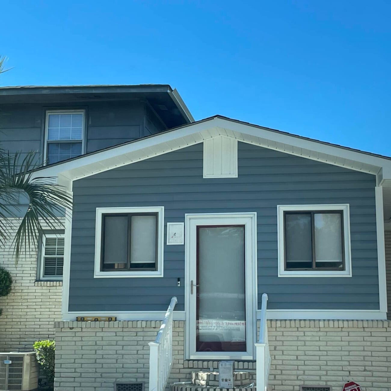 A small, single-story house with expert siding installation features blue-gray siding, white trim, brick foundation, and two front windows, sitting next to a larger two-story home under a clear blue sky.